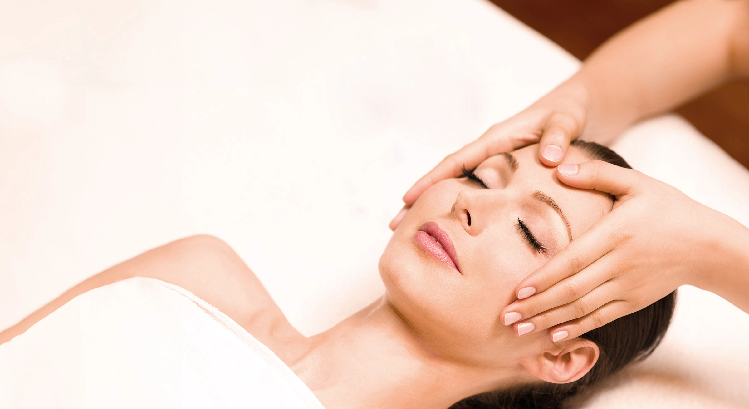 Entspannte Frau mit geschlossenen Augen liegt auf einer Massageliege und erhält eine sanfte Gesichtsmassage. Sie ist in ein weißes Handtuch gehüllt, der Hintergrund ist weich und hell gehalten, was eine ruhige Wellness-Atmosphäre vermittelt.