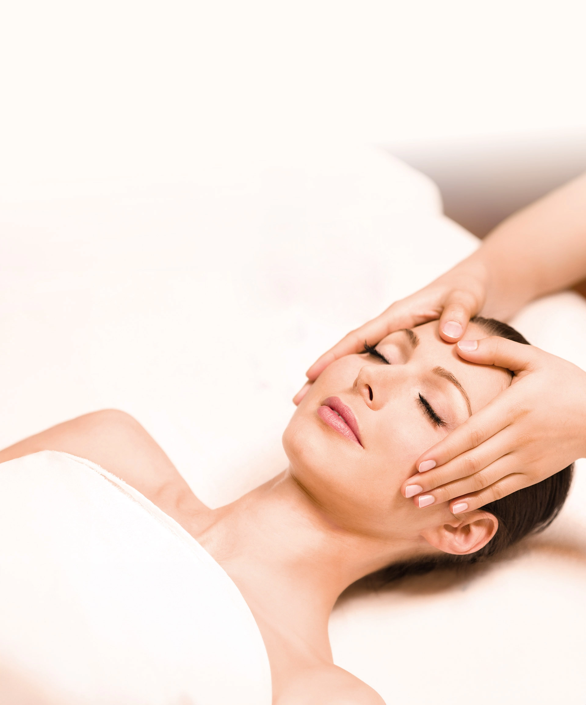 Entspannte Frau mit geschlossenen Augen liegt auf einer Massageliege und erhält eine sanfte Gesichtsmassage. Sie ist in ein weißes Handtuch gehüllt, der Hintergrund ist weich und hell gehalten, was eine ruhige Wellness-Atmosphäre vermittelt.